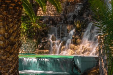 Yunanistan 'ın Loutraki kentinde taş duvarlar ve palmiyelerle çevrili sakin bir şelale. Yumuşak su hareketleri ve doğal tonlar rahatlama görselleri ya da turizm için mükemmeldir..