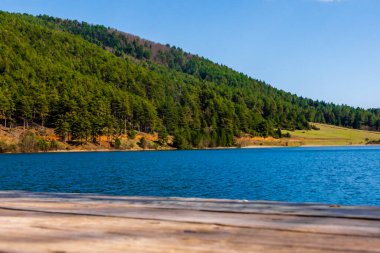Yunanistan 'ın Corinth kentindeki Doxa Gölü' ne bakan ahşap bir rıhtımdan manzara, turkuvaz suları, çam kaplı dağlar ve güneşli bahar gökyüzü kusursuz kaçışı..