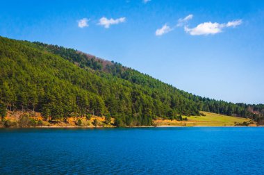 Yunanistan 'ın Corinth kentindeki Doxa Gölü, açık mavi suları, yemyeşil ormanları ve güneşli bir bahar gününde gökyüzünde birkaç bulutla yakalanan manzaralı dağ yamaçları..