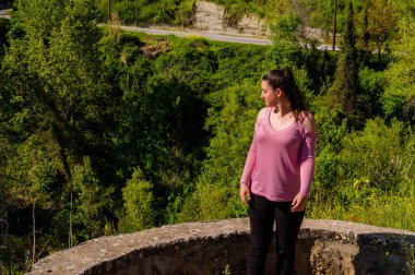 Genç bir kadın, Trikala, Corinth, Yunanistan 'da canlı bahar yeşiliyle çevrili pembe bir gömlek giyerek dışarıda duruyor. Doğal ışık ve huzurlu ruh hali..