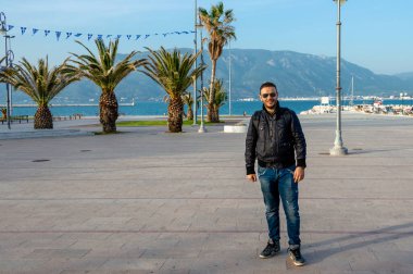 Yunanistan 'ın Corinth kentinin sahil şeridinde dikilen adamın arkasında palmiye ağaçları, Yunan bayrakları, deniz ve dağlar var. Öğleden sonra güneşinin altında rahat bir poz.