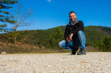 Yunanistan 'ın Corinth kentindeki Doxa Gölü yakınlarında yeşil orman tepeleri ve açık mavi gökyüzü ile çevrili kayalık kırsal bir yolda bir adam poz veriyor..