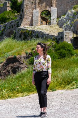 Genç bir kadın, güneşli bir bahar gününde, Yunanistan 'ın Corinth kentindeki Acrocorinth kalesinin ortaçağ taş kapısının önünde güven içinde duruyor..