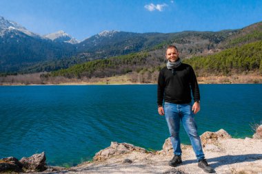 Doxa Gölü, Corinth, Yunanistan 'ın yanında duran bir adamın portresi, derin mavi sular, çam ormanı ve parlak mavi gökyüzünün altındaki uzak dağ zirveleriyle çevrili..