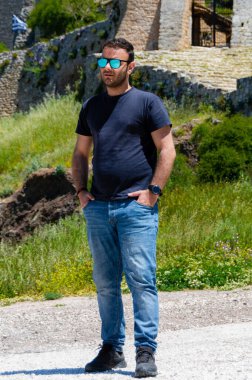 Yunanistan 'ın Corinth kentindeki Acrocorinth kalesinin önünde duran bir adamın gündelik portresi, arka planda bir bahar günü parlak güneş ışığı altında Yunan bayrağı.