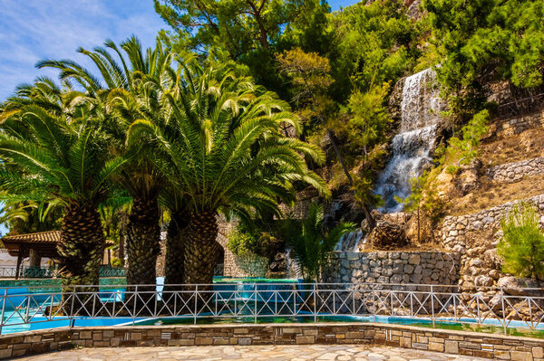 A scenic park in Loutraki, Corinth, Greece with tropical palm trees, a beautiful artificial waterfall and clear blue sky. Perfect Mediterranean summer scene.