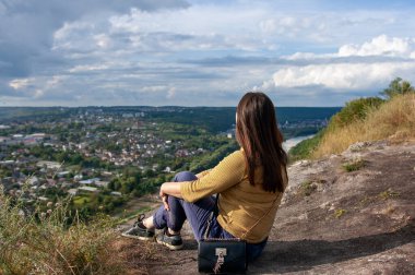 Genç bir kadın kayalık bir tepede tek başına oturmuş aşağıdaki vadiyi nefes kesen panoramik manzaraya bakıyor. Doğayla iç içe, tek başına seyahat etmenin özgürlüğünü ve huzurunu kucaklıyor..