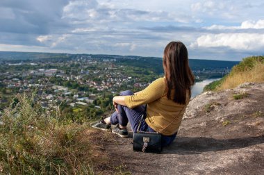 Genç bir kadın kayalık bir tepede tek başına oturmuş aşağıdaki vadiyi nefes kesen panoramik manzaraya bakıyor. Doğayla iç içe, tek başına seyahat etmenin özgürlüğünü ve huzurunu kucaklıyor..