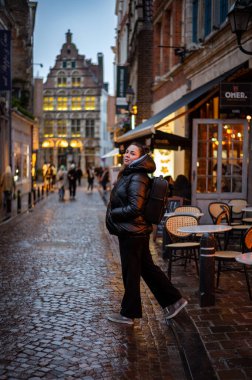 Tek başına gezen biri, samimi kafeler ve tarihi binalarla dolu büyüleyici bir Avrupa caddesinde akşam yürüyüşünden zevk alıyor. Işıkların ılık parıltısı ıslak kaldırım taşına yansıyarak büyülü bir atmosfer yaratıyor..