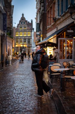 Tek başına gezen biri, samimi kafeler ve tarihi binalarla dolu büyüleyici bir Avrupa caddesinde akşam yürüyüşünden zevk alıyor. Işıkların ılık parıltısı ıslak kaldırım taşına yansıyarak büyülü bir atmosfer yaratıyor..