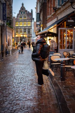Tek başına gezen biri, samimi kafeler ve tarihi binalarla dolu büyüleyici bir Avrupa caddesinde akşam yürüyüşünden zevk alıyor. Işıkların ılık parıltısı ıslak kaldırım taşına yansıyarak büyülü bir atmosfer yaratıyor..