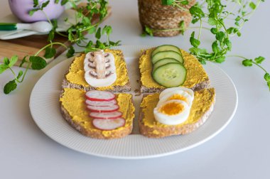 Kaynamış yumurta, salatalık, turp ve mantar gibi farklı malzemelerle kaplanmış dört açık yüzlü sandviç beyaz bir mutfakta beyaz bir tabakta servis edilir..