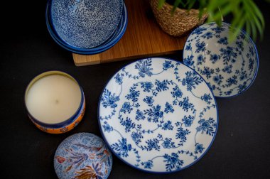 Güzel desenli mavi ve beyaz porselen kaseler ve tabaklar, bir saksı bitkisi ve dekoratif bir mum eşliğinde, rafine bir sofra takımı oluşturur.