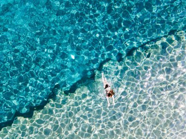 Canlı mavi suyun yüzeyinde yatan bir kızın büyüleyici bir hava görüntüsü. Vücudu ve sonsuz genişlikteki su arasındaki zıtlık çarpıcı ve minimalist bir kompozisyon oluşturuyor.