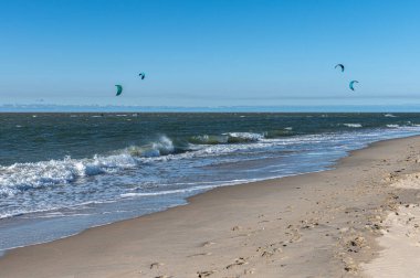 Uçurtma noktasında koruyucu giysiler içinde dalgalara binen birden fazla uçurtmacının panoramik görüntüsü. Aydınlık bir gün ve meltem bu adrenalin dolu su sporu macerası için mükemmel bir ortam yaratır..