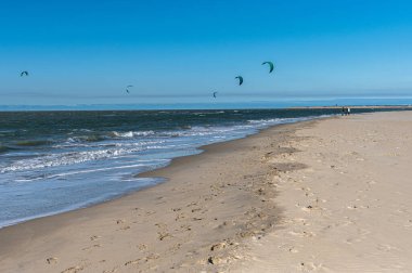 Uçurtma noktasında koruyucu giysiler içinde dalgalara binen birden fazla uçurtmacının panoramik görüntüsü. Aydınlık bir gün ve meltem bu adrenalin dolu su sporu macerası için mükemmel bir ortam yaratır..
