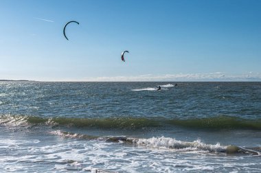 Uçurtma noktasında koruyucu giysiler içinde dalgalara binen birden fazla uçurtmacının panoramik görüntüsü. Aydınlık bir gün ve meltem bu adrenalin dolu su sporu macerası için mükemmel bir ortam yaratır..