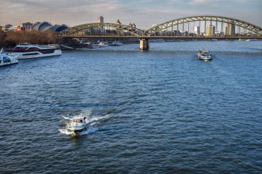 Rhein nehrinin üzerindeki Köln manzarası ve arka planda Hohenzollern Köprüsü. Nehirde iki araç.
