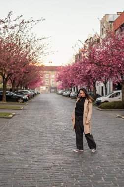 Belçika, Gent 'te bir kaldırım taşı caddesinde çiçek açan kiraz ağaçlarının altında bej ceketli bir kadının büyüleyici bir sahnesi. Yumuşak pembe yapraklar rüya gibi bir bahar havası yaratır..