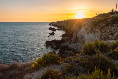Maltas 'ın nefes kesici manzarası engebeli taştan kıyı şeridi ılık günbatımı renkleriyle yıkanmış, kayalardan yansıyan altın ışık ve sakin Akdeniz.