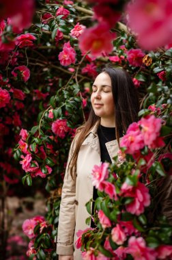 Genç bir kadın, gür kamelya çalılarıyla çevrili, canlı yapraklara daldırılmış. Narin çiçekler bahar hafifliği ve doğal uyum ortamı yaratır.