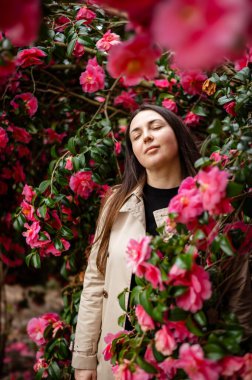 Genç bir kadın, gür kamelya çalılarıyla çevrili, canlı yapraklara daldırılmış. Narin çiçekler bahar hafifliği ve doğal uyum ortamı yaratır.