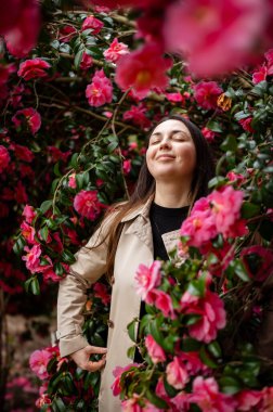 Genç bir kadın, gür kamelya çalılarıyla çevrili, canlı yapraklara daldırılmış. Narin çiçekler bahar hafifliği ve doğal uyum ortamı yaratır.