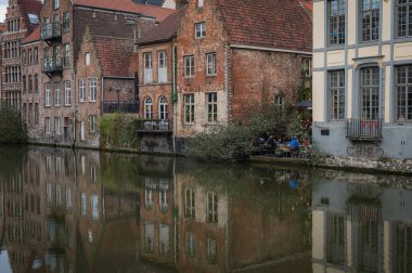 Gent, Belçika - 23 Mart 2025: Belçika 'nın Gent kentinin muhteşem tarihi mimarisi ve su üzerindeki yansımaları ile çevrili, kanallar boyunca yürüyen insanları gösteren fotografik bir çekimi.
