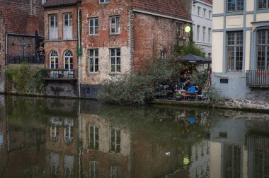 Gent, Belçika - 23 Mart 2025: Belçika 'nın Gent kentinin muhteşem tarihi mimarisi ve su üzerindeki yansımaları ile çevrili, kanallar boyunca yürüyen insanları gösteren fotografik bir çekimi.