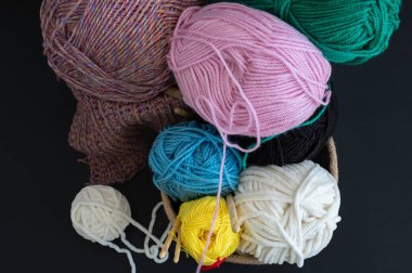 Örülmüş sepetteki renkli iplik toplarından oluşan bir koleksiyon, yanında da siyah arka planda beyaz bir iplik yumağı var. Örgü meraklıları ve el işleri için mükemmel.