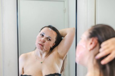 Neşeli, yarı çıplak, parlak ciltli bir kadının yakın çekimi. Nazikçe gülerken, rutin cilt kreminin bir parçası olarak nemlendirici yüz kremi sürüyor.