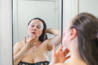 Neşeli, yarı çıplak, parlak ciltli bir kadının yakın çekimi. Nazikçe gülerken, rutin cilt kreminin bir parçası olarak nemlendirici yüz kremi sürüyor.