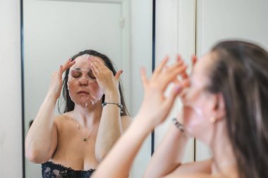 Neşeli, yarı çıplak, parlak ciltli bir kadının yakın çekimi. Nazikçe gülerken, rutin cilt kreminin bir parçası olarak nemlendirici yüz kremi sürüyor.