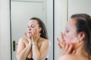 Neşeli, yarı çıplak, parlak ciltli bir kadının yakın çekimi. Nazikçe gülerken, rutin cilt kreminin bir parçası olarak nemlendirici yüz kremi sürüyor.