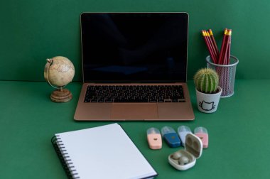 Yeşil bir arkaplanda düzenli olarak dizilmiş not defterleri, kalemler, keçeli kalemler ve makaslar da dahil olmak üzere okul malzemelerinden oluşan canlı bir çeşitlilik.