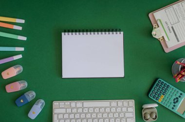 Yeşil bir arkaplanda düzenli olarak dizilmiş not defterleri, kalemler, keçeli kalemler ve makaslar da dahil olmak üzere okul malzemelerinden oluşan canlı bir çeşitlilik.