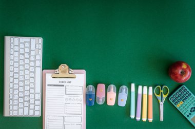 Yeşil bir arkaplanda düzenli olarak dizilmiş not defterleri, kalemler, keçeli kalemler ve makaslar da dahil olmak üzere okul malzemelerinden oluşan canlı bir çeşitlilik.