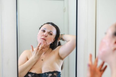 Neşeli, yarı çıplak, parlak ciltli bir kadının yakın çekimi. Nazikçe gülerken, rutin cilt kreminin bir parçası olarak nemlendirici yüz kremi sürüyor.