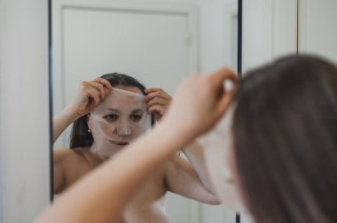 Neşeli, yarı çıplak, parlak ciltli bir kadının yakın çekimi. Nazikçe gülerken, rutin cilt kreminin bir parçası olarak nemlendirici yüz kremi sürüyor.