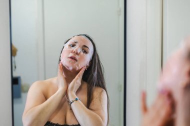 Neşeli, yarı çıplak, parlak ciltli bir kadının yakın çekimi. Nazikçe gülerken, rutin cilt kreminin bir parçası olarak nemlendirici yüz kremi sürüyor.