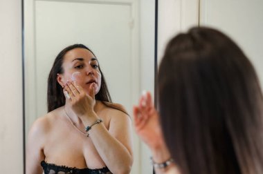 Neşeli, yarı çıplak, parlak ciltli bir kadının yakın çekimi. Nazikçe gülerken, rutin cilt kreminin bir parçası olarak nemlendirici yüz kremi sürüyor.