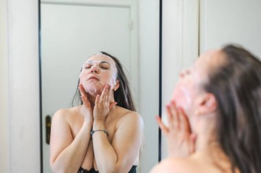 Neşeli, yarı çıplak, parlak ciltli bir kadının yakın çekimi. Nazikçe gülerken, rutin cilt kreminin bir parçası olarak nemlendirici yüz kremi sürüyor.