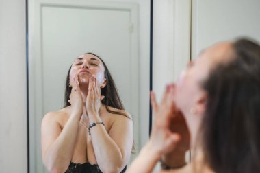 Neşeli, yarı çıplak, parlak ciltli bir kadının yakın çekimi. Nazikçe gülerken, rutin cilt kreminin bir parçası olarak nemlendirici yüz kremi sürüyor.