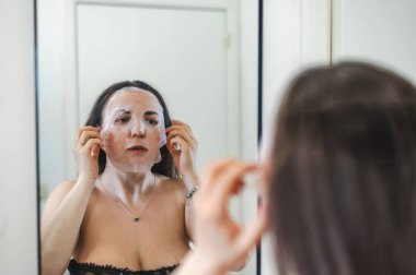 Neşeli, yarı çıplak, parlak ciltli bir kadının yakın çekimi. Nazikçe gülerken, rutin cilt kreminin bir parçası olarak nemlendirici yüz kremi sürüyor.