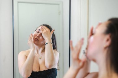 Neşeli, yarı çıplak, parlak ciltli bir kadının yakın çekimi. Nazikçe gülerken, rutin cilt kreminin bir parçası olarak nemlendirici yüz kremi sürüyor.