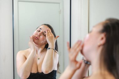 Neşeli, yarı çıplak, parlak ciltli bir kadının yakın çekimi. Nazikçe gülerken, rutin cilt kreminin bir parçası olarak nemlendirici yüz kremi sürüyor.