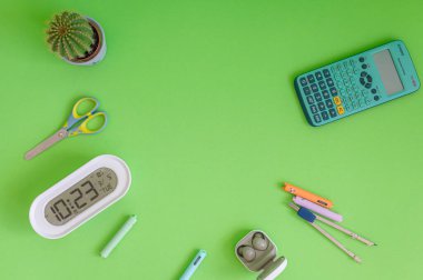 Yeşil bir arkaplanda düzenli olarak dizilmiş not defterleri, kalemler, keçeli kalemler ve makaslar da dahil olmak üzere okul malzemelerinden oluşan canlı bir çeşitlilik.