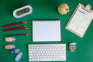 Yeşil bir arkaplanda düzenli olarak dizilmiş not defterleri, kalemler, keçeli kalemler ve makaslar da dahil olmak üzere okul malzemelerinden oluşan canlı bir çeşitlilik.