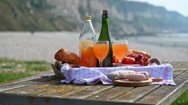 Normandiya 'da Fransız cilası, bölgesel peynir ve taze elmaların yer aldığı samimi bir plaj pikniği manzaralı bir sahilin arka planında geçiyor.