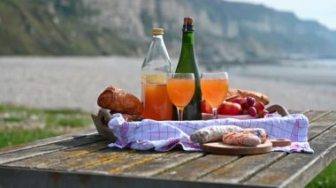 Normandiya 'da Fransız cilası, bölgesel peynir ve taze elmaların yer aldığı samimi bir plaj pikniği manzaralı bir sahilin arka planında geçiyor.